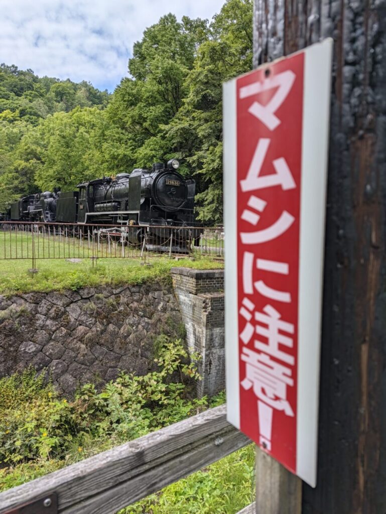 「マムシに注意」と書かれた赤い看板。奥に保存展示の蒸気機関車が見える